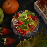 Colorful Fruit Jellies in a glass bowl with an orange, strawberry, and leaf on a dark surface.
