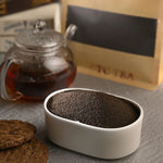 Tea powder in a white container with a glass teapot and cookies on a wooden surface.